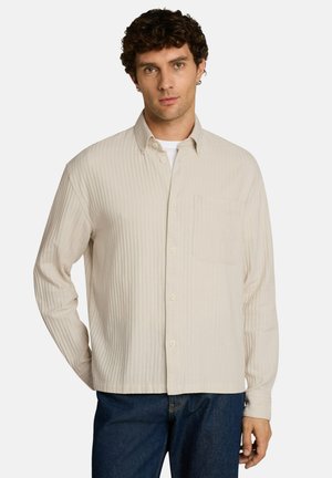 Man met krullend haar die een beige overhemd met knopen draagt over een wit T-shirt en donkerblauwe spijkerbroek, staand tegen een effen achtergrond.