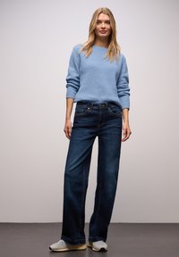 Jasnoniebieski sweter z dzianiny, ciemnoniebieskie spodnie dzwony, szare trampki z żółtymi podeszwami; gładka faktura, casualowy design i luźny krój.