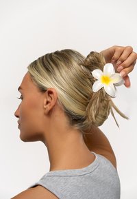 Plastiktillbehör för håret i form av en vit blomma med gul mitt, fäst i en knut av vågigt blont hår. Grå ärmlös topp synlig.