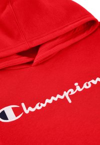 Felpa rossa con cappuccio realizzata in tessuto morbido, caratterizzata da un logo "Champion" bianco scritto in corsivo e un distintivo patch a forma di "C."
