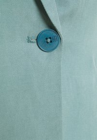 Jasnozielony blezer z teksturowanej tkaniny, z wyraźnym, okrągłym niebieskim guzikiem z czterema dziurkami i delikatnym połyskiem.