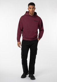 Burgunderfarbenes Hoodie mit Fronttasche, gesticktem "WORLDWIDE"-Logo, kombiniert mit schwarzen Cargo-Hosen und schwarzen Sneakers. Glatte Textur und lockere Passform.