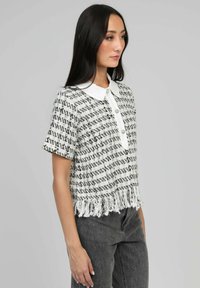 Haut court à manches courtes avec un col blanc, motif tissé noir et blanc, et détails à franges sur l'ourlet. Fermeture à trois boutons à l'avant.