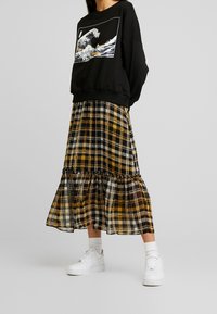 Sweat-shirt noir avec un imprimé graphique, associé à une jupe à carreaux jaune, noire et blanche, avec un ourlet à volants, et des baskets blanches.