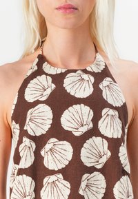 Smeđa halter top majica s uzorkom bijelih školjki; izrađena od lagane tkanine s teksturiranim završetkom i podesivim vezicom oko vrata.
