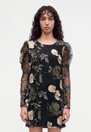 Femme aux longs cheveux bouclés portant une robe noire avec des manches bouffantes transparentes et une broderie florale beige, debout devant un fond clair uni.