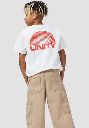 Ragazzo con dreadlocks con punte bionde che indossa una maglietta bianca con un semicerchio a strisce rosse e la scritta "UNITY", e pantaloni cargo beige, di profilo.