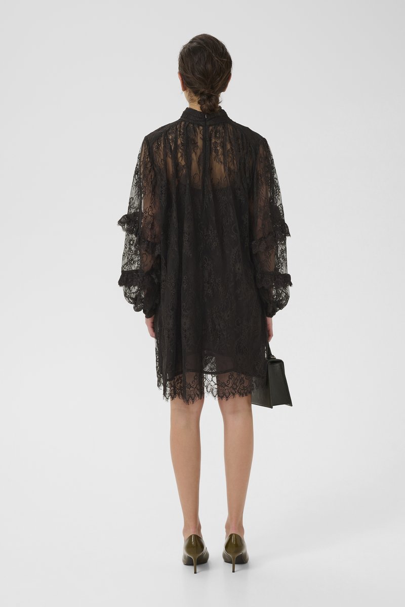 Black lace dress with ruffled sleeves, sheer design, and scalloped hem. Paired with a small green handbag and olive heels.