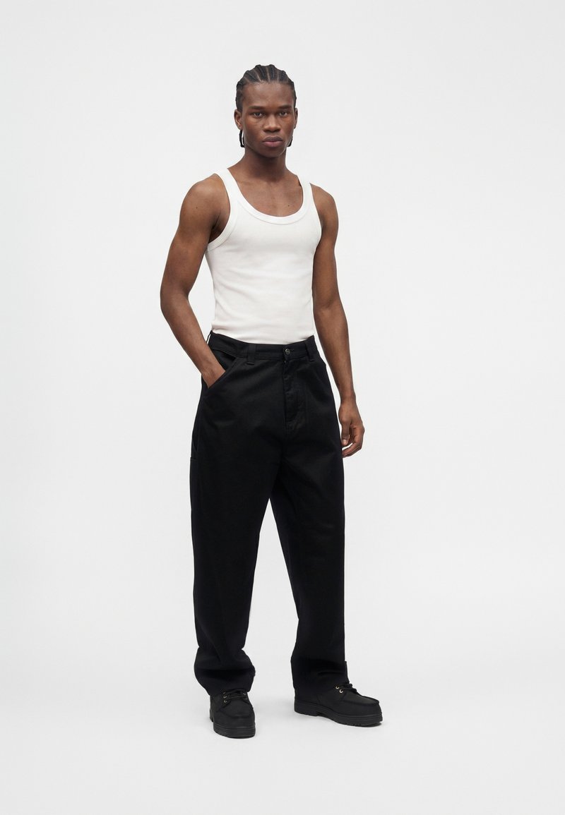 Young man with braided hair wearing a white sleeveless tank top, black loose pants, and black shoes standing with one hand in pocket.