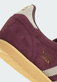 Μπορντό σουέτ αθλητικό παπούτσι με γκρι ακροβατικά, χρυσό λογότυπο και υφή σόλας από καουτσούκ σε χρώμα καραμέλας με ζιγκ-ζαγκ μοτίβο.