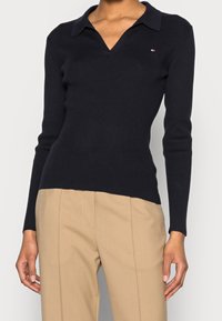 Maglione nero a coste con scollo a V e colletto. Caratterizzato da maniche lunghe e un piccolo logo sulla parte superiore sinistra.