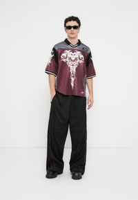 Chemise bordeaux à manches courtes avec motif graphique blanc et accents gris, associée à un pantalon noir ample avec des bandes latérales rouges et des chaussures noires épaisses.