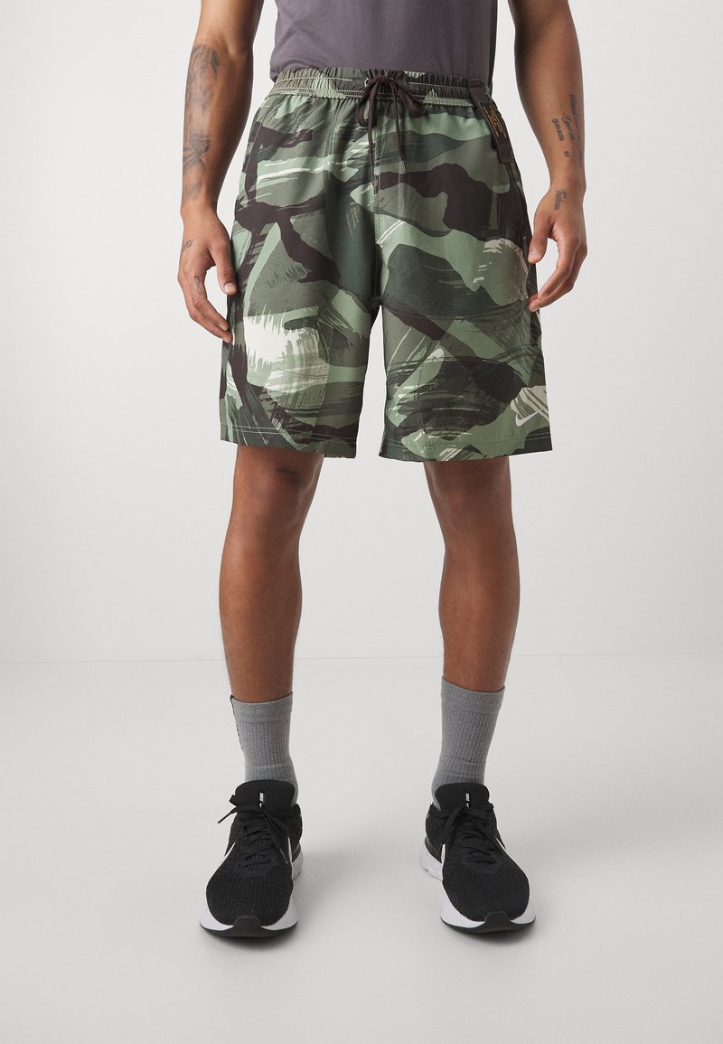 Mand iført grønne og sorte camouflage-shorts, grå sokker og sorte sportssko, stående mod en ensfarvet baggrund.