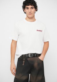 Λευκή βαμβακερή μπλούζα με κόκκινο γραφικό "Double" στο στήθος, συνδυασμένη με μαύρο παντελόνι με σκισμένο υφή και ζώνη με τρυπήματα.