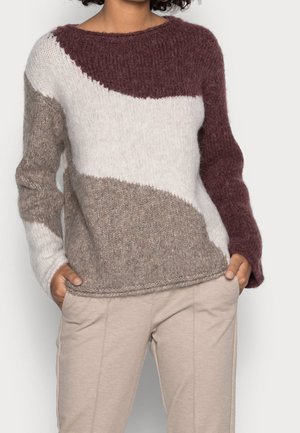 Femme portant un pull à manches longues bicolore en bordeaux, blanc et beige, les mains dans les poches d'un pantalon beige, debout devant un fond uni.