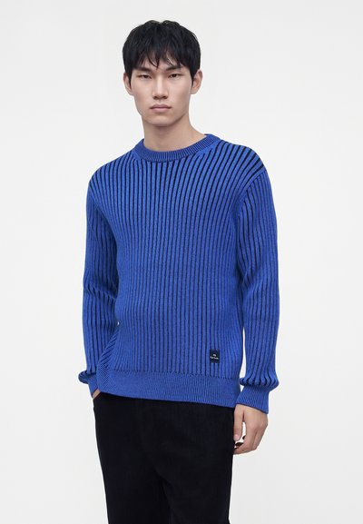 Jeune homme aux cheveux courts noirs, portant un pull en maille côtelée bleu et un pantalon noir, debout devant un fond blanc uni.