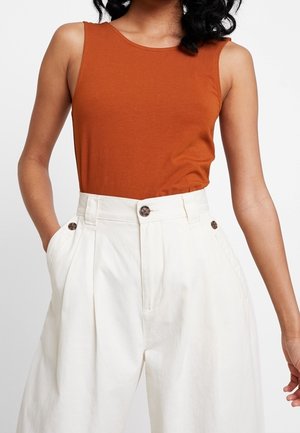 Kobieta w bezrękawniku w kolorze rdzawego pomarańczu, wpuszczonym w spodnie z wysokim stanem o szerokich nogawkach w kolorze białym, z ozdobnymi guzikami i rękami w kieszeniach.