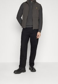 Grå ærmeløs vest over en ribbet grå sweater, parret med sorte bukser og sorte støvler med kraftig sål. Gul logo på vesten.