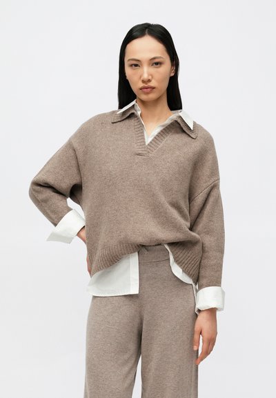 Pull en maille beige oversize avec col en V et ourlet côtelé, superposé à une chemise blanche à col. Texture douce et coupe décontractée.