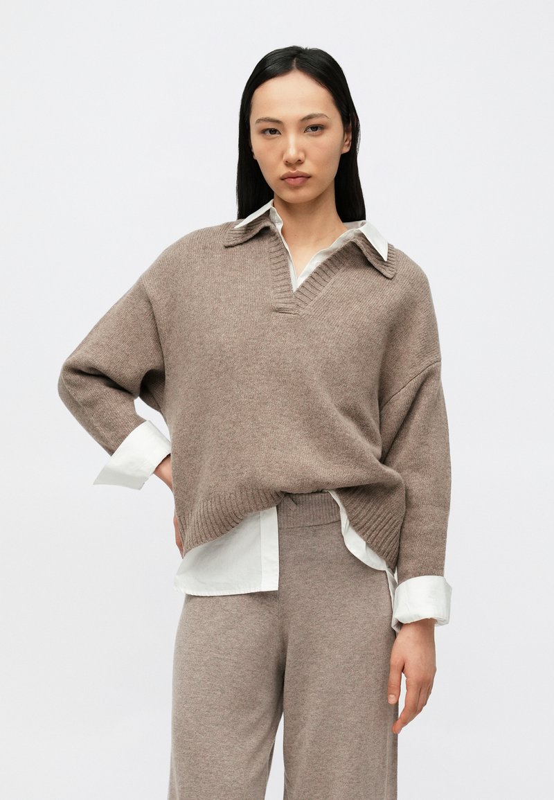 Beežikas, üle mõõdu kootud kampsun, millel on V-kaelus ja soonikkääris, kantud valge kraega särgi üle. Pehme tekstuur ja lõdvestunud lõige.