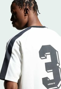 Balts sporta krekls ar melnām akcentiem, tajā skaitā liels melns numurs 3 uz aizmugures, ribotā apkakle un īsas piedurknes. Redzams Adidas logotips.