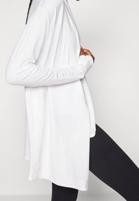 Top bianco a maniche lunghe con una vestibilità rilassata, texture liscia e spacco laterali, abbinato a leggings neri. Presenta un colletto rotondo.