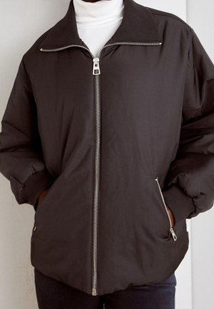 Tmavě hnědá bunda na zip s žebrovanými manžetami a límcem, nošená přes bílý rolák, ruce v zipovaných bočních kapsách.