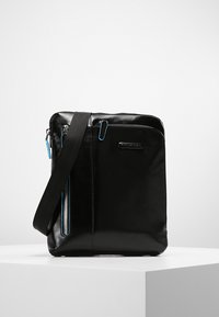 Bolsa mensajera de cuero negro con textura suave, bolsillo frontal con cremallera y tiradores de cremallera azul en contraste. Incluye una correa de hombro desmontable.