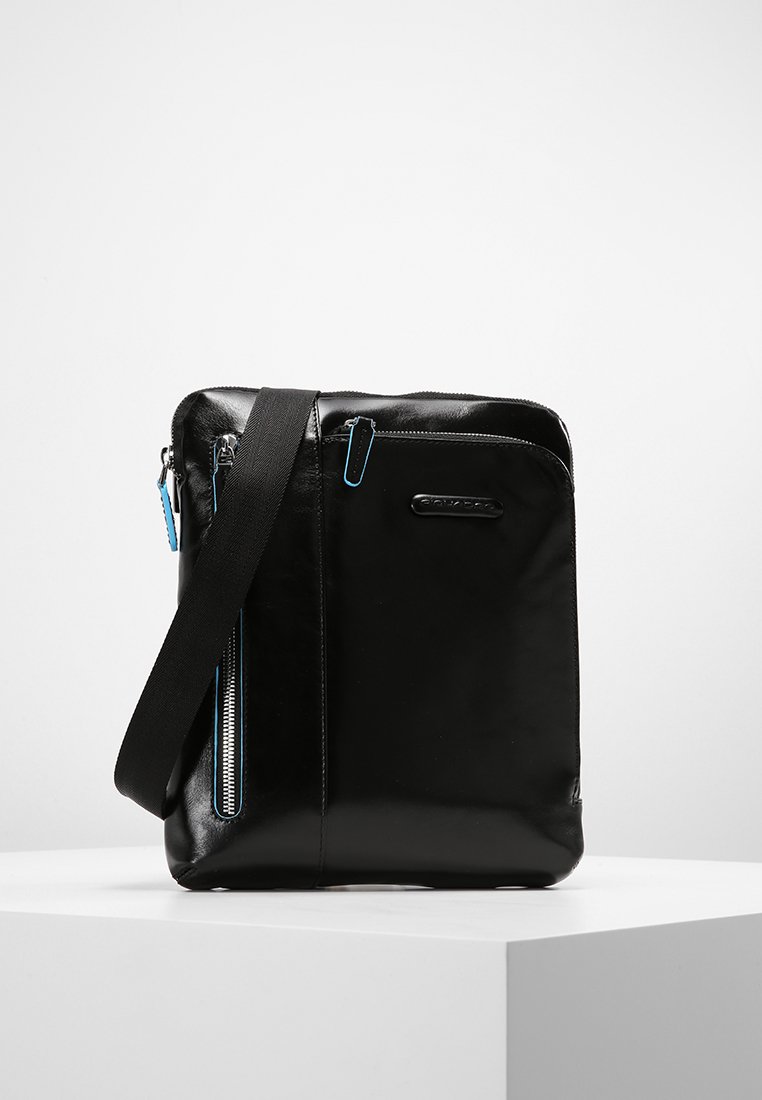 Bolsa mensajera de cuero negro con textura suave, bolsillo frontal con cremallera y tiradores de cremallera azul en contraste. Incluye una correa de hombro desmontable.