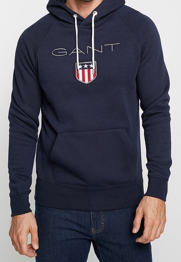 Un uomo indossa un hoodie blu navy con lacci bianchi, una tasca frontale e il testo "GANT" sopra un emblema con uno scudo rosso, bianco e blu.