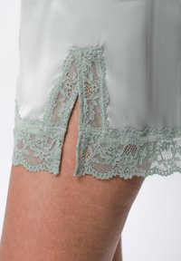 Primo piano di un tessuto verde pallido con bordi in pizzo, caratterizzato da un motivo floreale e dettagli di spacco, indossato su pelle nuda nella parte superiore della coscia.