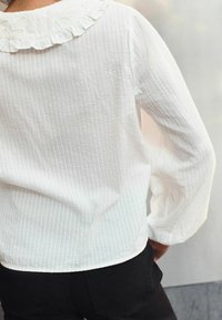 Weiße, strukturierte Bluse mit vertikalen Streifen, ausgestattet mit einem Rüschkragen und langen Ärmeln, kombiniert mit schwarzen, eng anliegenden Hosen.