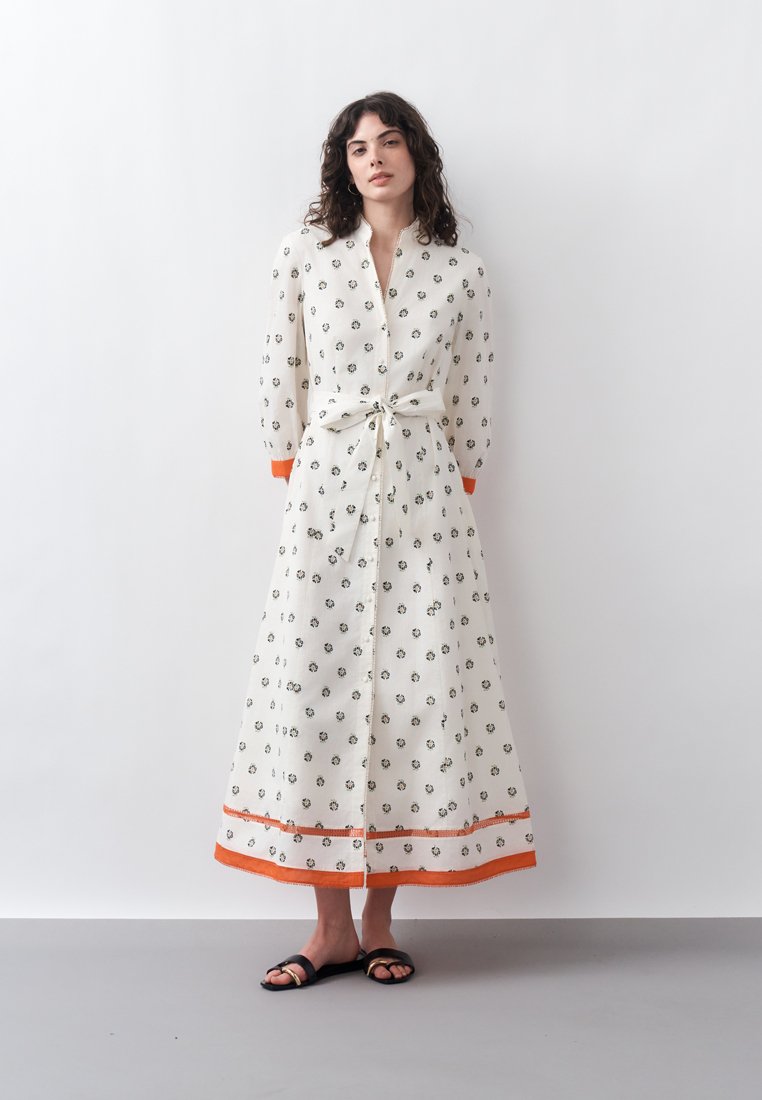 Femme debout contre un mur blanc, portant une longue robe blanche avec de petits motifs floraux, bordure orange, ceinture nouée et sandales noires.