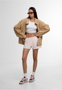 Veste oversized beige, crop top blanc, shorts rose clair et baskets marron/roses. Les accessoires incluent des lunettes de soleil rectangulaires. Textures lisses.
