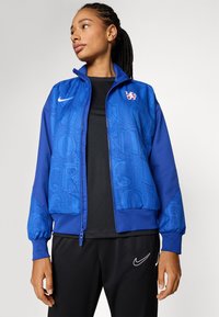 Plava zip-up jakna s reljefnim uzorkom teksta, rebrastim rukavima i Nike logom. Nosit će se preko crne majice, u kombinaciji s crnim hlačama.