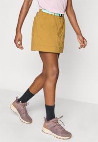 Παντελόνι σορτς χακί με υφή, που διαθέτει τιρκουάζ ζώνη και δύο πλαϊνές τσέπες. Συνδυάζεται με γκρι και εκρού παπούτσια πεζοπορίας και μαύρες κάλτσες.