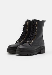 Musta nahkasaappaat, joissa on paksut kulutuspinnat, kultaiset lenkit, mustat nauhat ja metallinen "H"-riipus etunauhoissa.