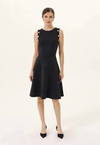 Vestido preto sem mangas com corpete ajustado e saia rodada. Apresenta botões dourados nos ombros e uma textura suave. Usado com sapatos pretos de bico.
