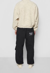 Giacca pullover beige pelosa, taglio ampio con orlo arrotondato. Pantaloni cargo neri con tasche, con testo logo bianco sulla coscia sinistra.