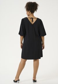Robe noire courte avec des manches bouffantes et un dos en V, dotée d'une fermeture à nouer. Texture lisse et coupe décontractée, se terminant au-dessus du genou.