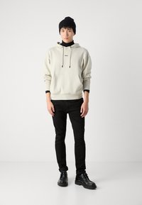 Sudadera crema con cordones y bolsillo frontal, combinada con pantalones negros y botines negros. Un gorro negro completa el look.