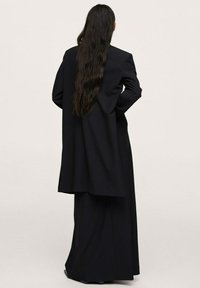 Zwarte op maat gemaakte jas met een lange achterkant, gedragen over een bijpassende jurk tot op de vloer. Heeft lange mouwen en een gladde textuur.