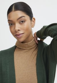 Cardigan verde a maglia sopra un collo alto marrone. Il cardigan presenta una vestibilità comoda e dettagli a coste sulle maniche. Tessuto liscio.