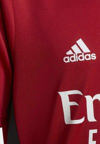 Camiseta deportiva roja de Adidas con el logo blanco de Adidas y texto parcial en blanco sobre una tela texturizada, mostrando detalles de las mangas y los paneles laterales.