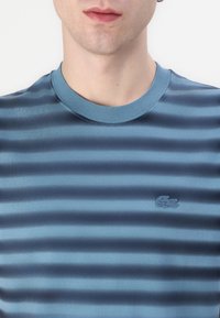 Niebieska t-shirt w paski z gładkiego materiału, z prążkowanym okrągłym dekoltem i wyhaftowanym logo w lewym dolnym rogu.