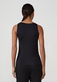Donna con capelli intrecciati, che indossa una canotta nera senza maniche e pantaloni neri, in piedi di fronte a uno sfondo grigio chiaro uniforme.