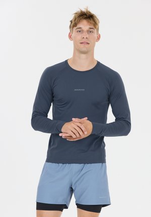 Junger Mann mit zerzausten Haaren, der ein dunkelblaues, langärmeliges Sportshirt und hellblaue Shorts trägt, vor einem schlichten weißen Hintergrund steht.
