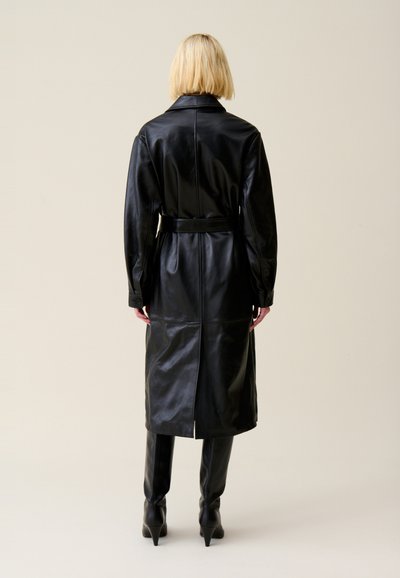 Long manteau en cuir noir avec une ceinture, doté d'un col à revers, de manches longues et d'une fente au dos. Texture lisse et design épuré.