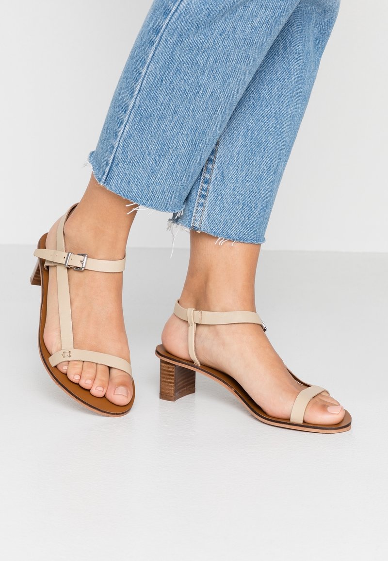 Beige sandaletter med låg klack. Har justerbara ankelremmar och en slät läderfinish. Bärs med avklippta blå jeans.