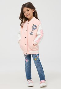 Giacca varsity rosa e bianca con toppa di unicorno e jeans blu con ricami colorati a tema spaziale. Sneakers bianche con fasce pastello.